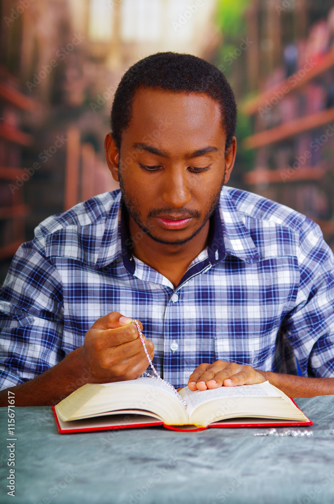 Religious man sitting while holding rosary, praying and reading from open book on desk in front