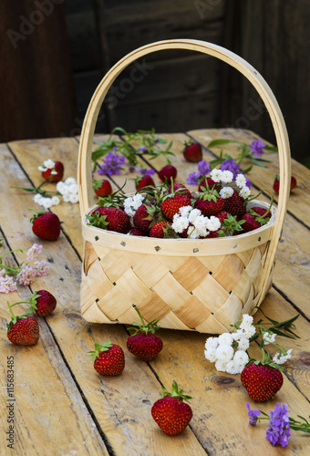 Bast basket with strawberries on a light wooden table with scattered berries and flowers 
