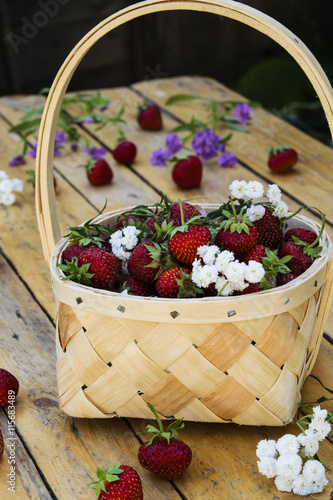 Bast basket with strawberries on a light wooden table with scattered berries and flowers 