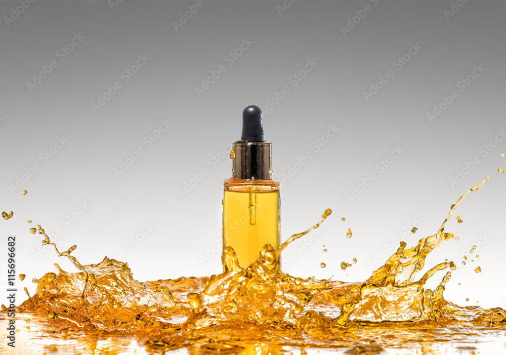 The bottle of the yellow cosmetic in the big orange oil splash on the gradient grey background The bottle of the yellow cosmetic in the big orange oil splash on the gradient grey background