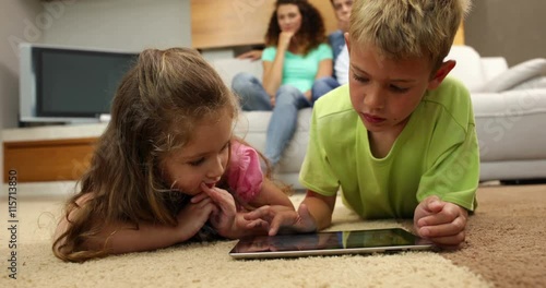 Sibings lying on floor using tablet with parents behind them on sofa photo