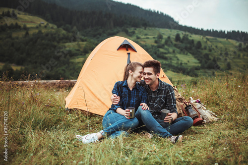 A couple of tourists in time of the hike, sit and relax outside the tent, admiring the beautiful mountain scenery. The guy hugs the girl. The concept of love, tenderness and recreation