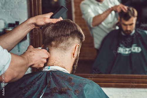 The hands of young barber making haircut to attractive man in barbershop