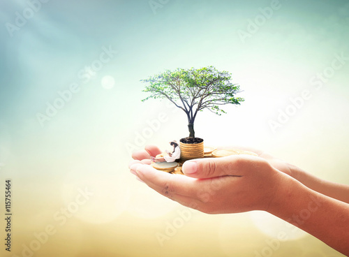 World environment day concept: Human hand holding a girl reading book, stack of golden coins and big tree over blurred nature background