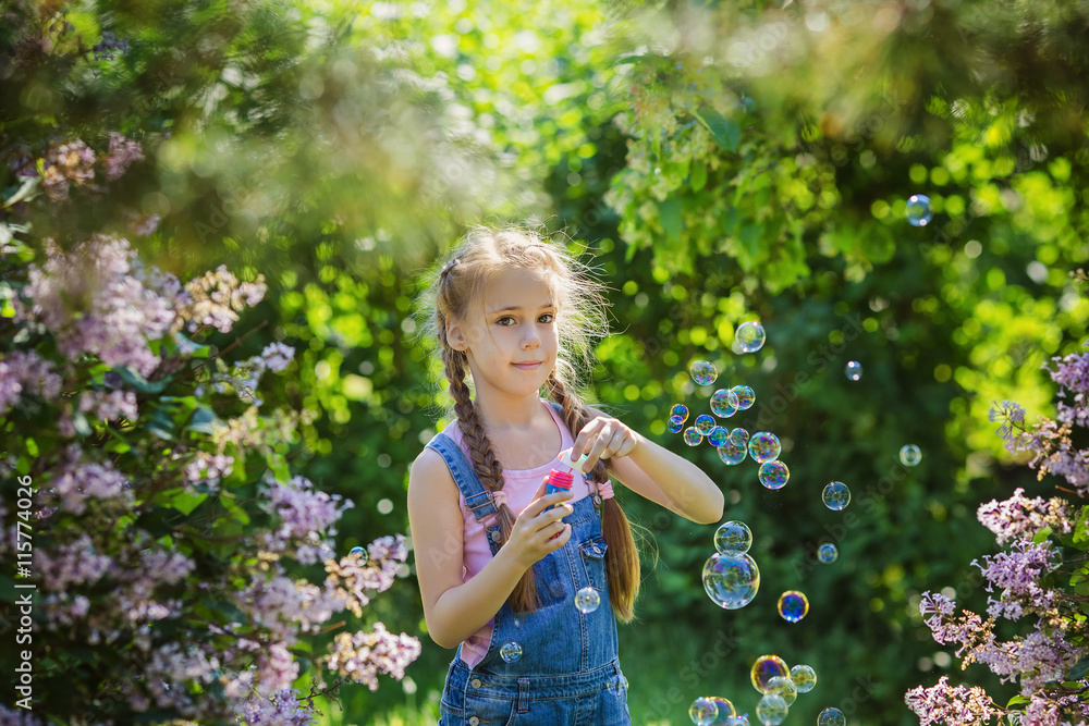 Beautiful little girl in denim overalls blowing bubbles in lilac blossoming garden