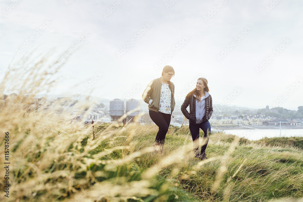 Happy female friends talking while walking on grassy hill against sky Happy female friends talking while walking on grassy hill against sky