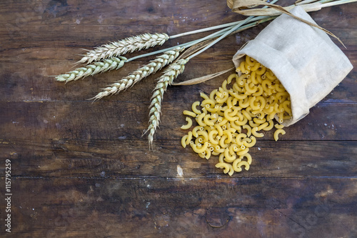 Uncooked Italian pasta Elbow macaroni in canvas little bag with wheat ears on dark wooden background.