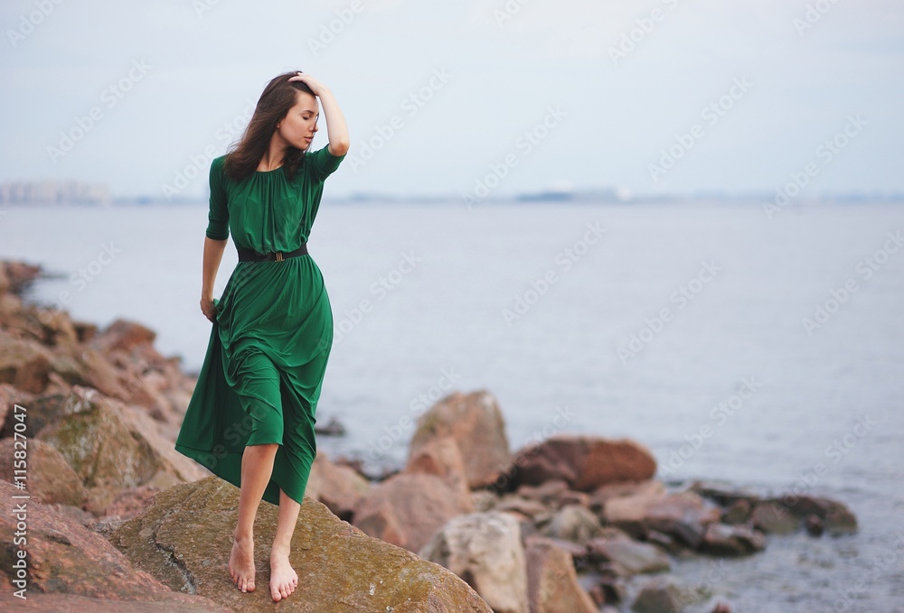 Beautiful slender girl in elegant Green Gown stands barefoot on the big rock on the shore of the Bay.