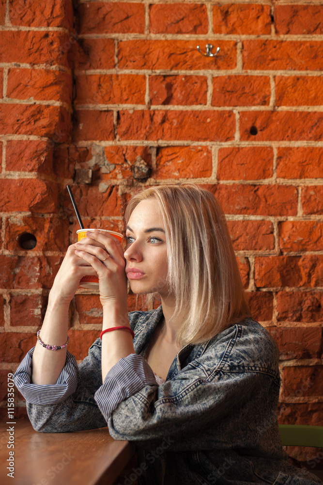 Young blonde woman holding orange paper cup with cocktail straw sitting near window against red brick wall at the cafe
