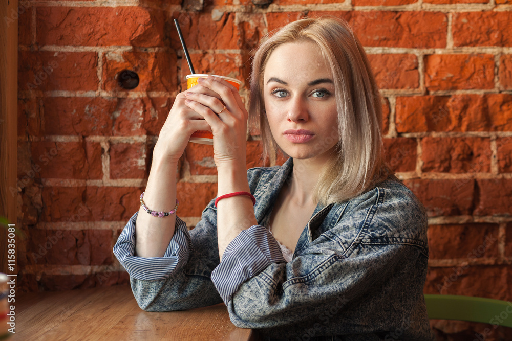Obraz premium Young blonde woman holding orange paper cup with cocktail straw sitting near window against red brick wall at the cafe quizzically looking at the camera
