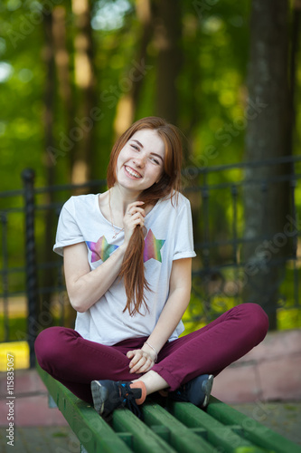 Young smiling beautiful redhead woman in red jeans sneakers shirt sitting on bench touching her hair blurry forest park background
