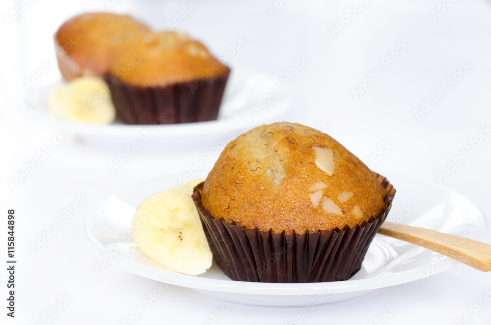 Close-up pieces of fresh delicious banana cup cakes topped with sliced almond in dark brown paper cup with fresh sliced sweet banana over white plate with wooden spoon are over white table background Close-up pieces of fresh delicious banana cup cakes topped with sliced almond in dark brown paper cup with fresh sliced sweet banana over white plate with wooden spoon are over white table background