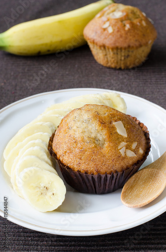 Two pieces of fresh delicious banana cup cake topped with sliced almond in dark brown paper cup with fresh sliced banana over white plate with wooden spoon was put over brown cloth napery.