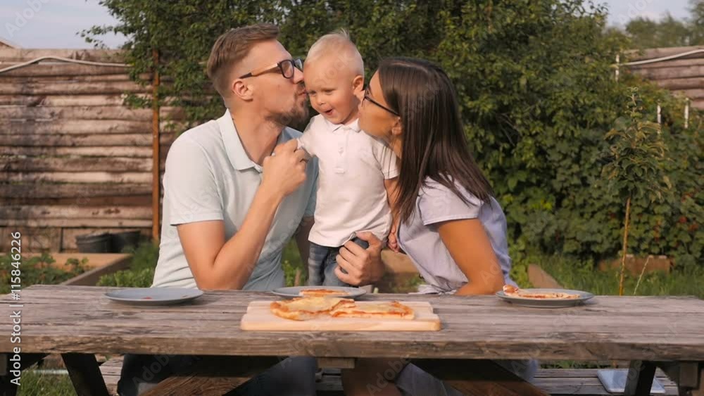 Young Happy Family Having Picnic In The Garden. Mom, Dad And Son Eating Pizza Outdoors Young Happy Family Having Picnic In The Garden. Mom, Dad And Son Eating Pizza Outdoors