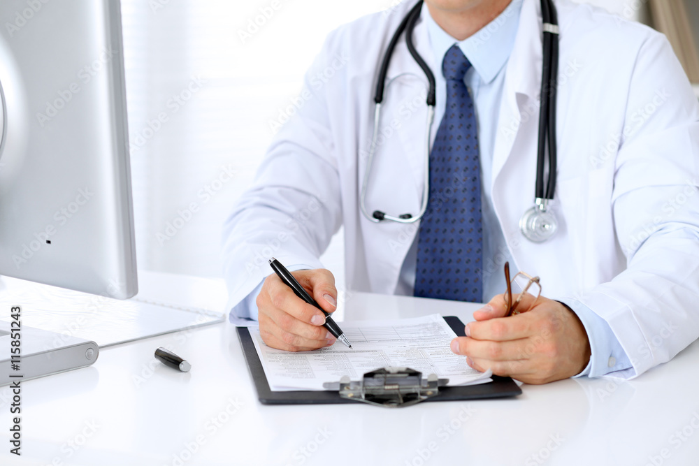 Close up of male doctor is sitting at the table and filling up medical history form Close up of male doctor is sitting at the table and filling up medical history form