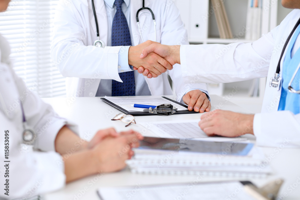 Two doctors shaking hands to each other sitting at the table in hospital office Two doctors shaking hands to each other sitting at the table in hospital office