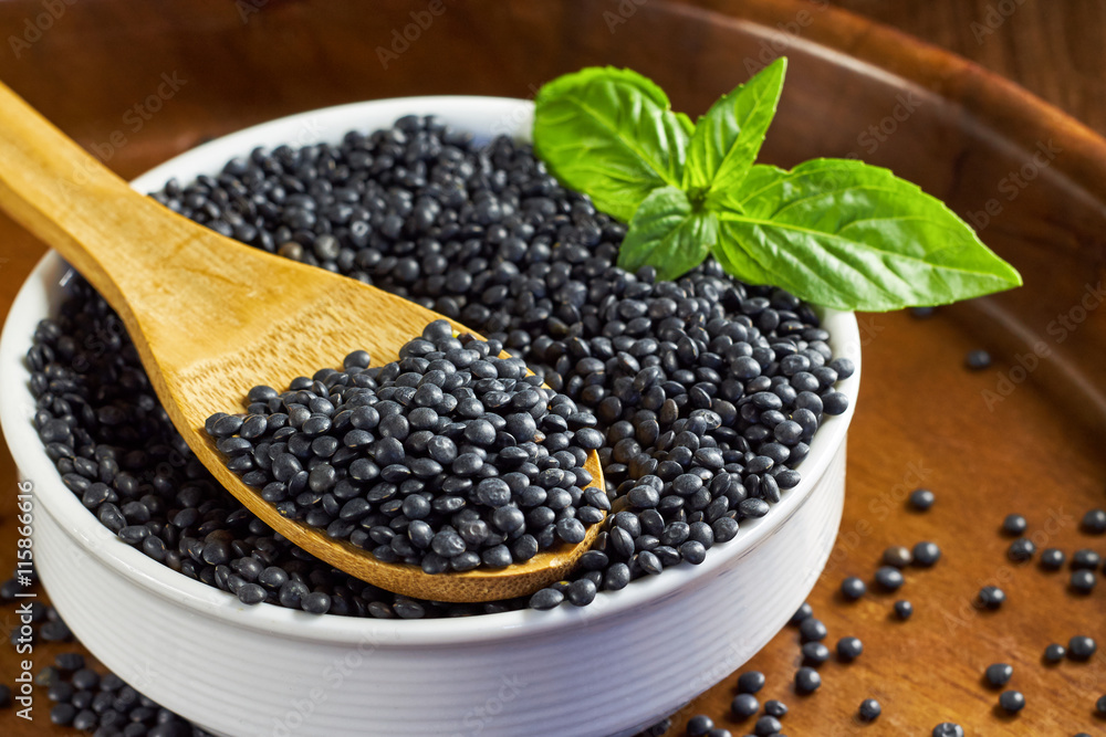 Round white bowl full of beluga lentils and basil leaves with wooden spoon on brown wooden background. Round white bowl full of beluga lentils and basil leaves with wooden spoon on brown wooden background.