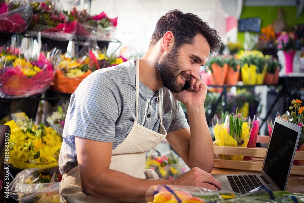 Naklejka premium Male florist talking on mobile phone while using laptop