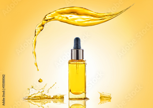The bottle of the yellow cosmetic oil with big splash above and many splashes around on the gradient yellow background