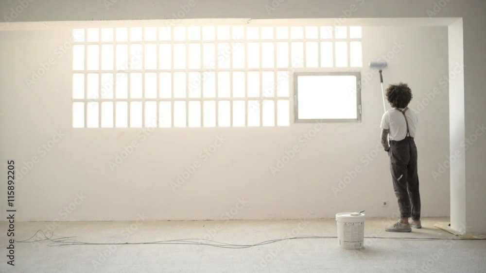 Funny curly man in pants with suspenders paints the white wall with brush roller near big industrial window Home diy renovation Loop Funny curly man in pants with suspenders paints the white wall with brush roller near big industrial window Home diy renovation Loop