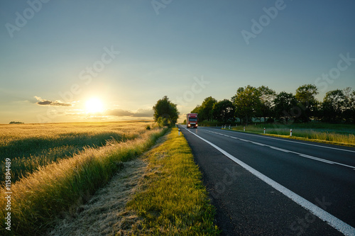 An asphalt road along the corn field at sunset. Red truck coming from afar.