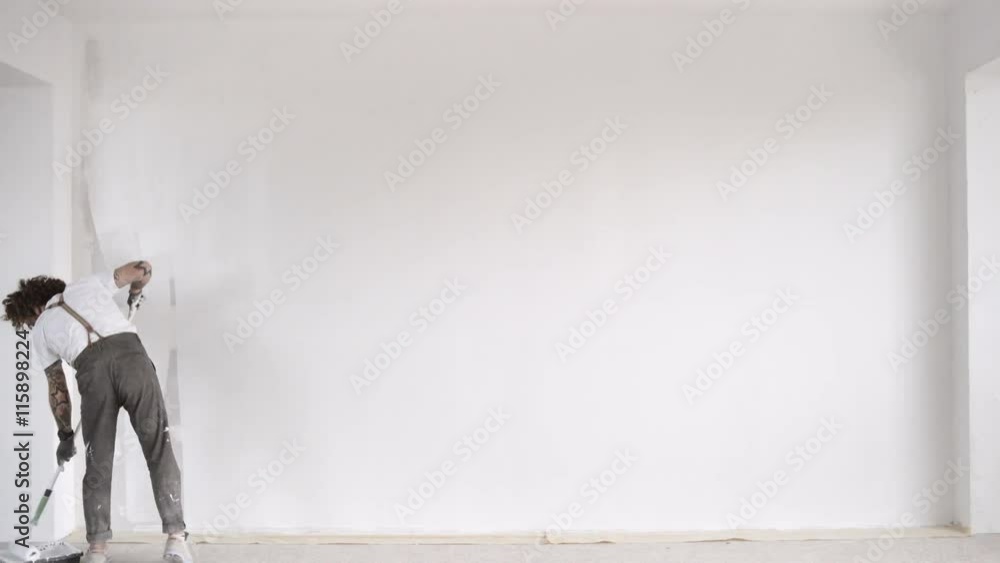Funny curly man wears pants with suspenders quickly finishes to paint big wall from plasterboard with white paint and brush roller Home dyi renovation Funny curly man wears pants with suspenders quickly finishes to paint big wall from plasterboard with white paint and brush roller Home dyi renovation