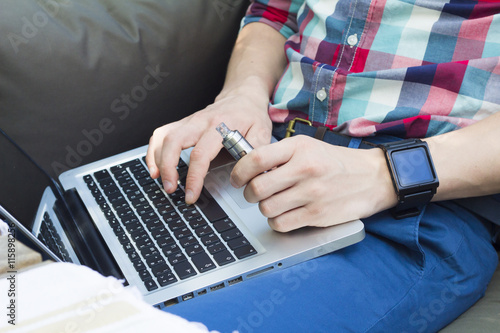 Cropped image of modern man typing something on laptop, with watch and e-cigarette.