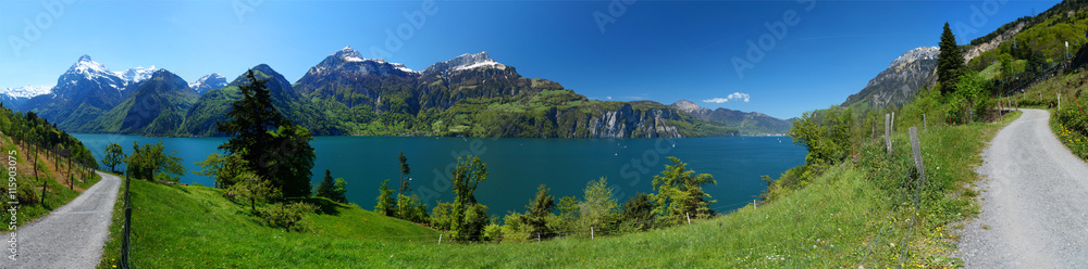 Hiking trail next to a calm mountain lake surrounded by snow covered peaks and lush, green meadows in the Swiss Central Alps. Lake Lucerne (Vierwaldstättersee), Swiss. May 2016. ~200° view angle.