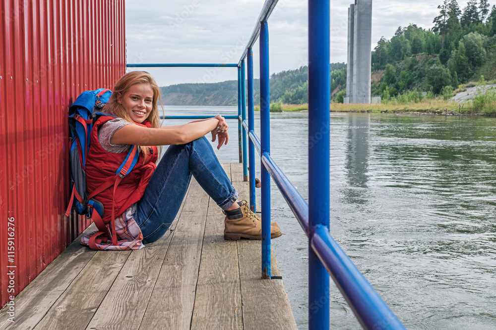 Blonde girl sitting on pier with backpack and smiling at camera
