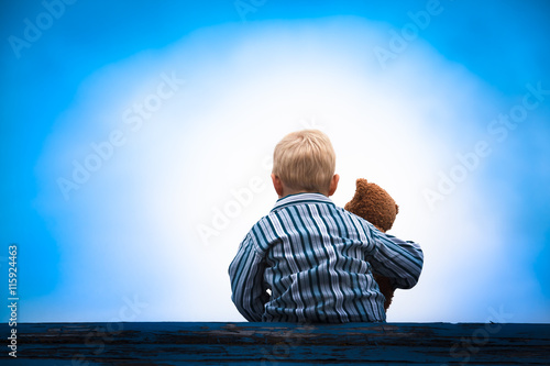 Child and Teddy at the Roof / Little boy in pajama cloth sitting at a wooden roof with the teddy bear in his arm in front of a bright light at the dark blue sky