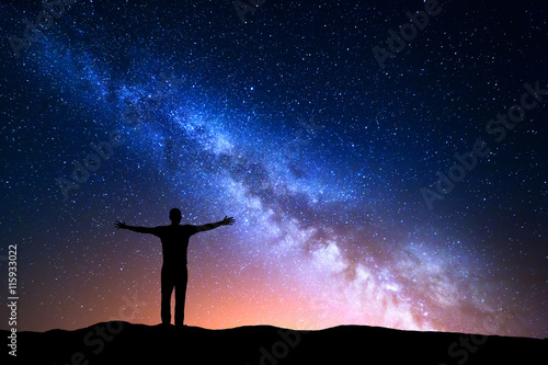 Nocny krajobraz z Drogą Mleczną. Sylwetka stojącego młodego człowieka z podniesionymi rękami na górze. Piękny wszechświat. Tło podróży z rozgwieżdżonym niebem niebieski noc