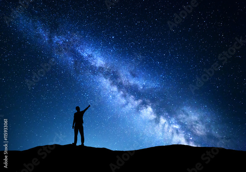 Milky Way. Silhouette of a standing man pointing finger in night starry sky on the mountain. Colorful night landscape with beautiful universe. Travel background with blue sky full of stars 