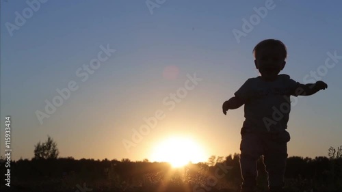 A little 1 year old baby boy is walking on sunset in nature. Child make his first steps outdoor. Kids Silhouette in the field with sundown