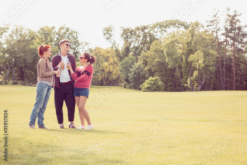 Image of happy best friends in sunglasses resting and relaxing in park. Cheerful people walking on green grass, communicating and drinking alcohol drinks.