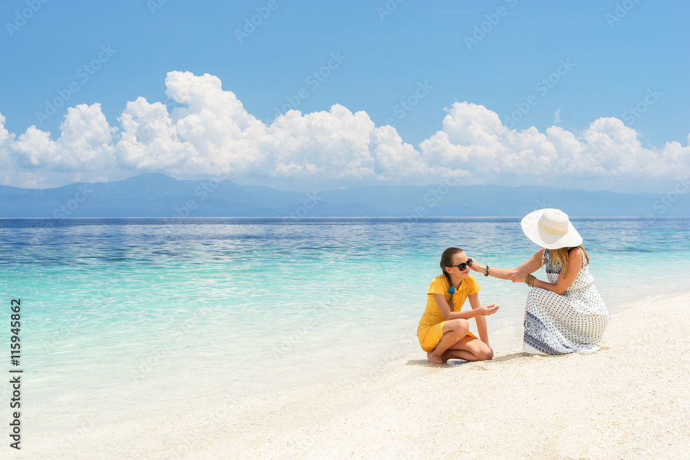 Youn europian woman in light dress is sitting on the white sand beach near beautiful tropical sea and cares about her teen daughter at sunny day