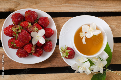 Top view of a mug of tea and jasmine flowers, a plate with strawberries on wooden box