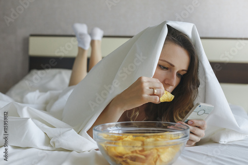 Girl eating chips in bed and watching news in phone