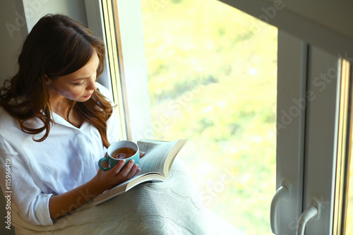 Young woman at home sitting near window relaxing in her living room reading book and drinking coffee or tea