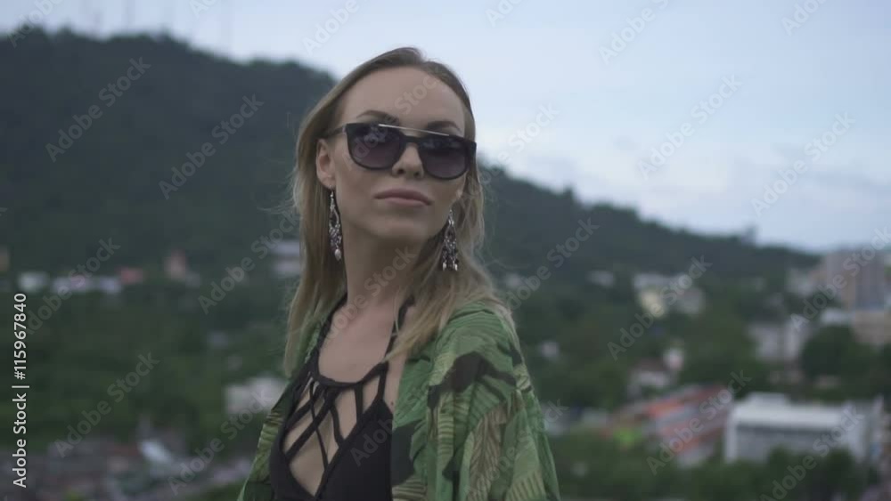 Sexy blonde woman wearing black swimwear and floral long shirt walking on a rooftop in a sunny summer day with beautiful sky, green mountain and citylandscape background - video in slow motion. Sexy blonde woman wearing black swimwear and floral long shirt walking on a rooftop in a sunny summer day with beautiful sky, green mountain and citylandscape background - video in slow motion.