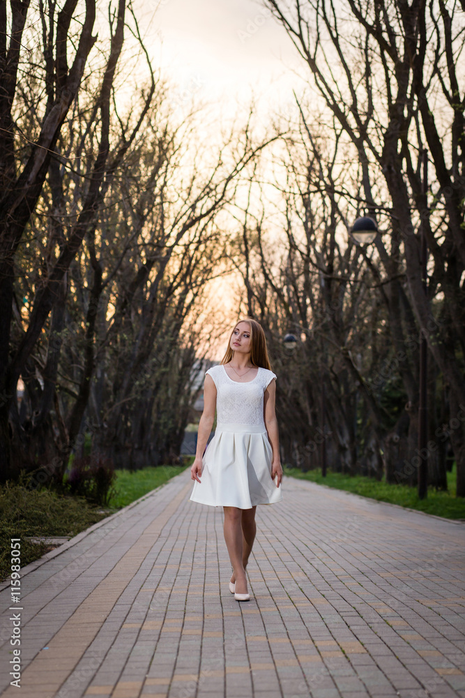 Beautiful blonde girl with long wavy curls in a white dress walk in evening the park among big trees.