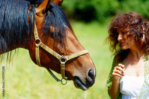 girl in white dress talking to a brown horse on meadow.