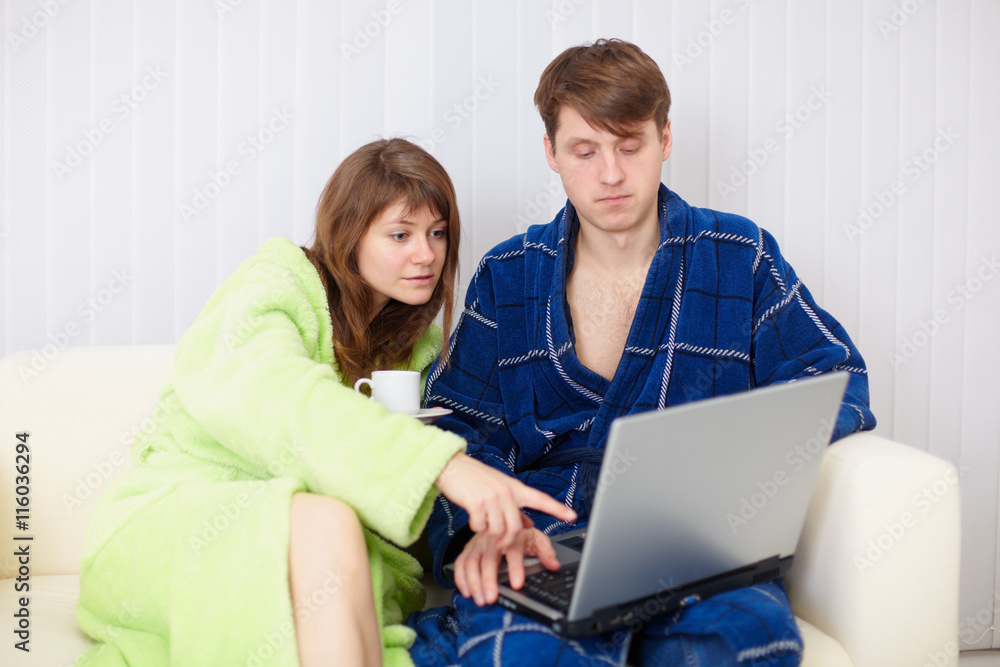 Spouses sit on sofa in dressing gowns with laptop Spouses sit on sofa in dressing gowns with laptop