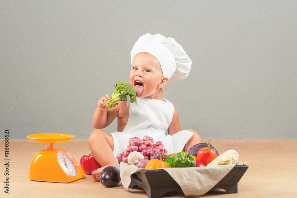 Little girl dressed in white chef hat and apron, eats only vegetables. The child weighed on the scales Products: eggplant, onion, garlic, pumpkin, red and green peppers, cabbage, tomatoes Little girl dressed in white chef hat and apron, eats only vegetables. The child weighed on the scales Products: eggplant, onion, garlic, pumpkin, red and green peppers, cabbage, tomatoes