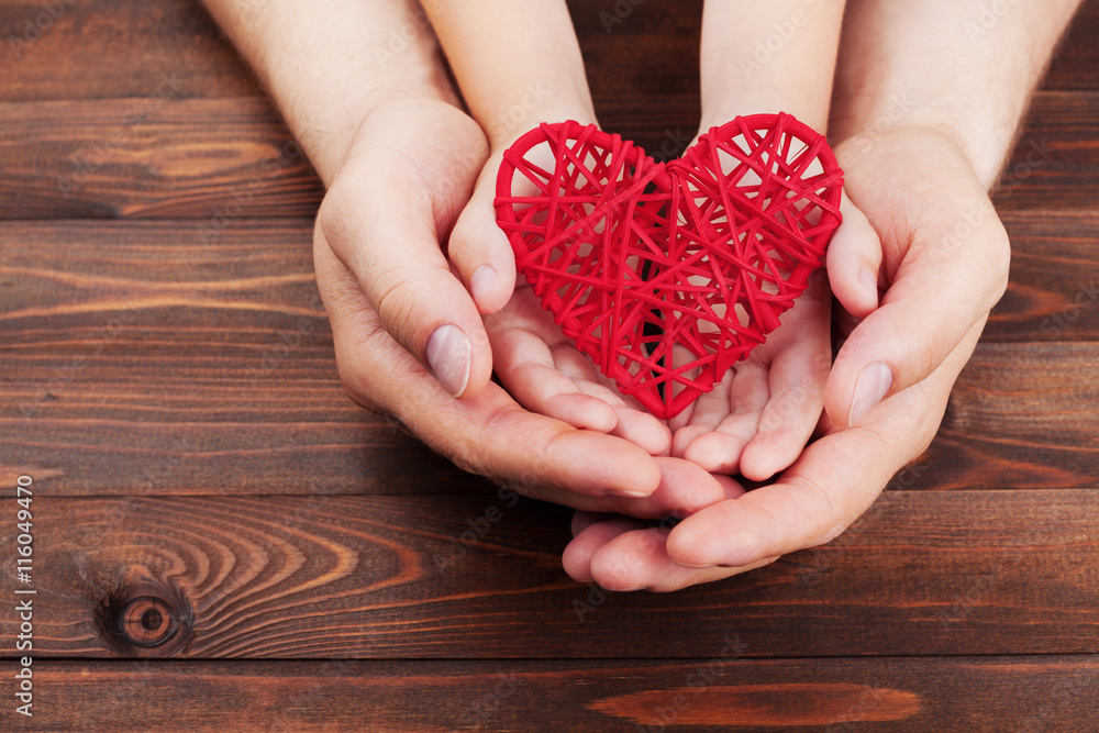 Adult and child holding red heart in hands over a wooden table. Family relationships, health care, pediatric cardiology concept. Adult and child holding red heart in hands over a wooden table. Family relationships, health care, pediatric cardiology concept.