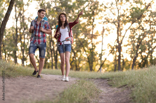 Young Couple Man and Girl in casual Travel Clothing with Backpacks walking on Forest Trail pointing with Hand toward Horizon