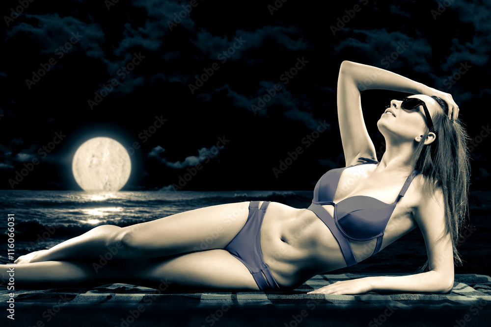 a lovely girl with long hair in a fashionable swimsuit lying on the beach in the moonlight in the dark background
