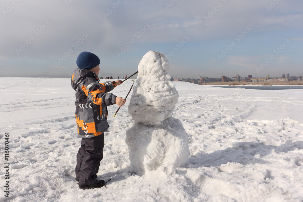 The little boy in a color jacket building a snowman on the river bank in the winter
