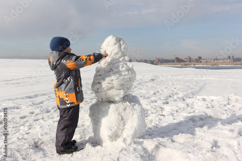 The little boy in a color jacket building a snowman on the river bank in the winter 