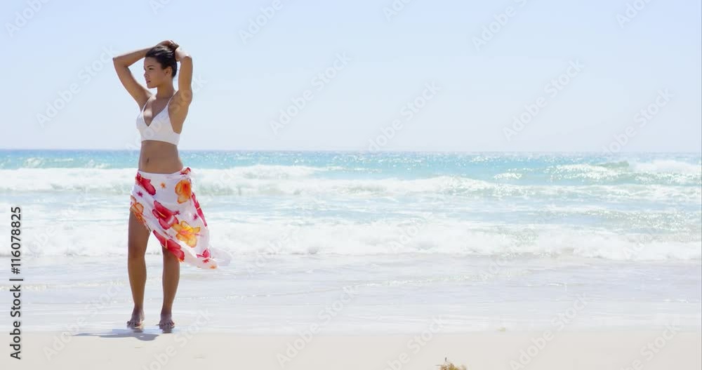 Gorgeous young woman walking along a beach in a bikini and colorful sarong at the waters edge on a summer day Gorgeous young woman walking along a beach in a bikini and colorful sarong at the waters edge on a summer day
