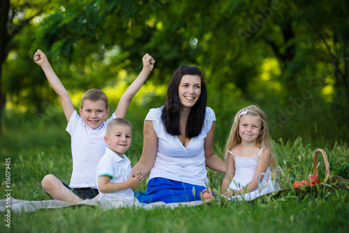Happy mother, two sons and daughter dressed in white shirts are sitting on the grass in the park